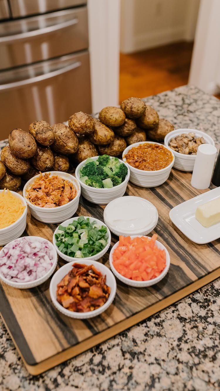 Baked potatoes and various toppings in white bowls on a wooden serving board.