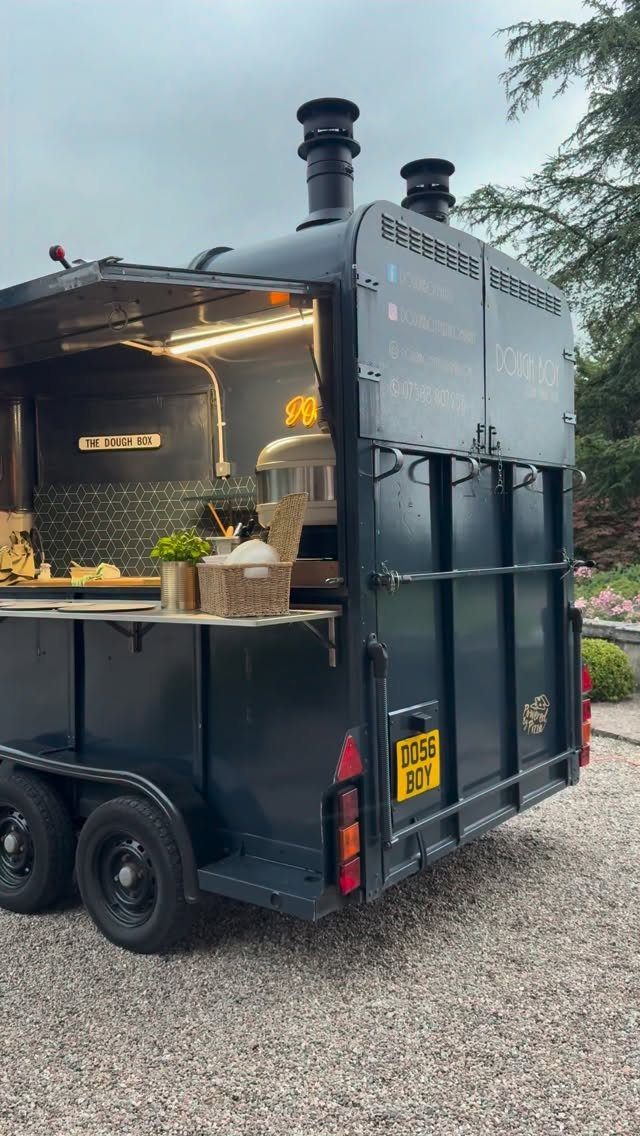 Black mobile food trailer with open serving hatch parked on a gravel driveway