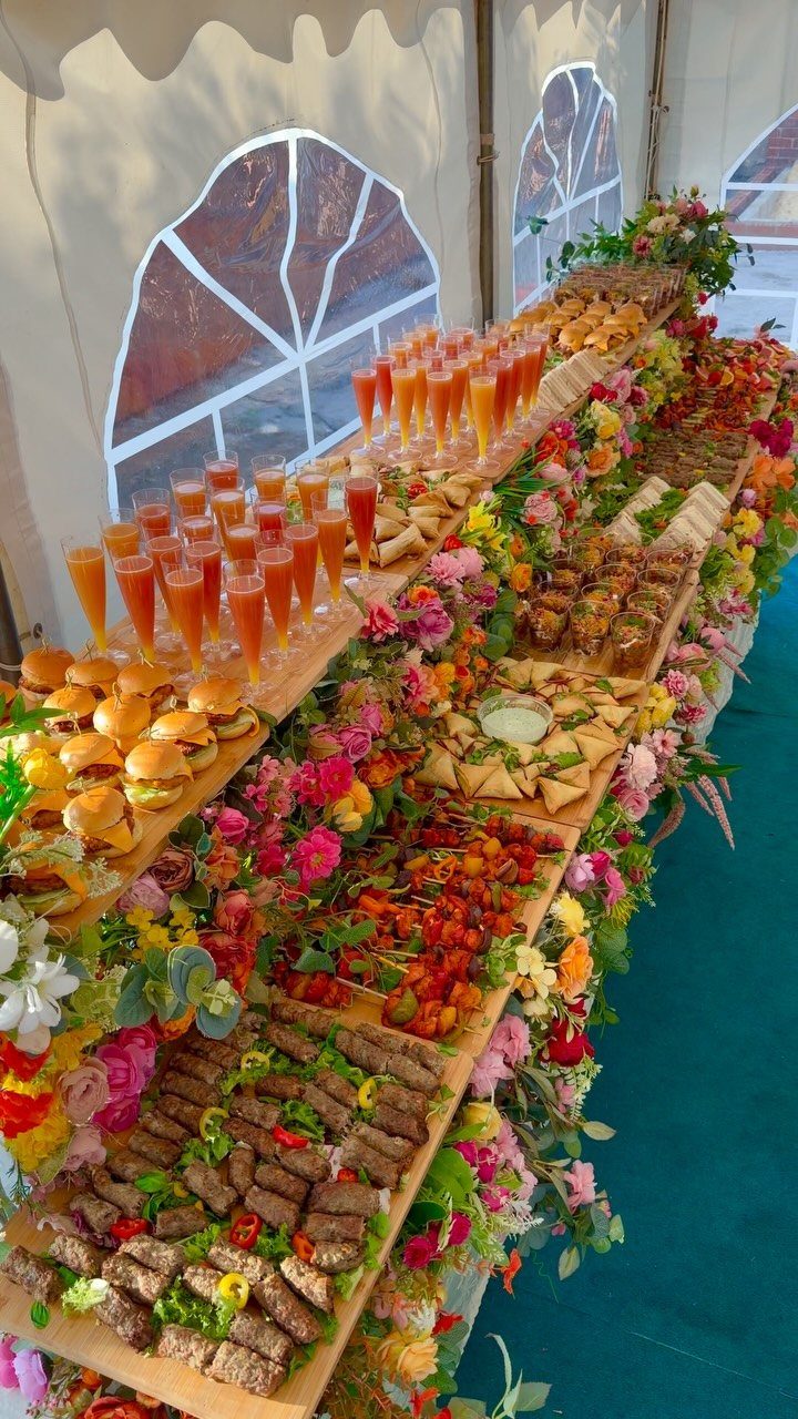 Multi-tiered wooden grazing table with sliders, drinks, and appetizers decorated with flowers.