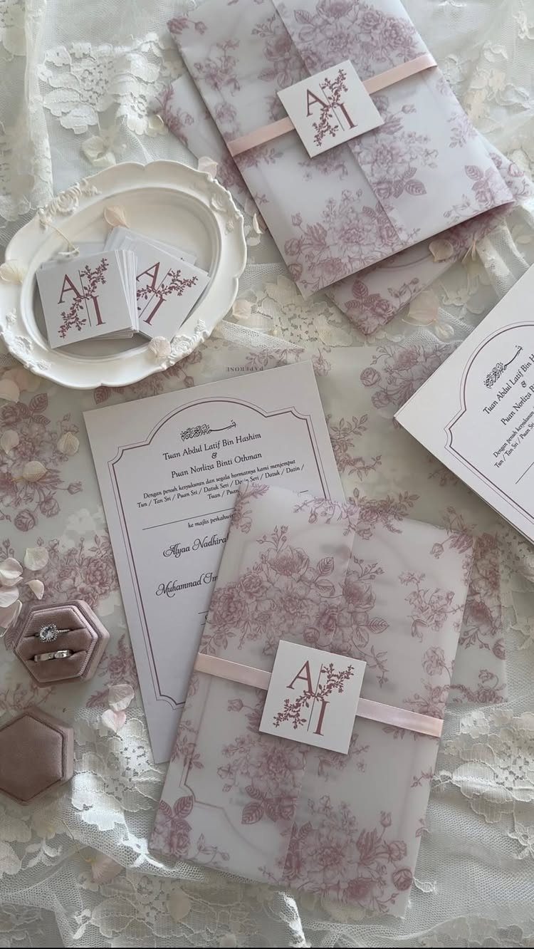 Floral purple wedding invitation envelopes with pink ribbons and monogrammed tags on lace.