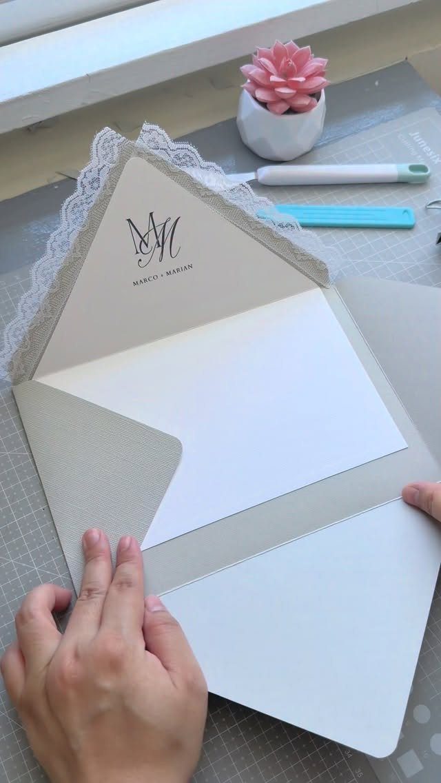 Beige wedding invitation envelope with white lace trim and a monogram being assembled.