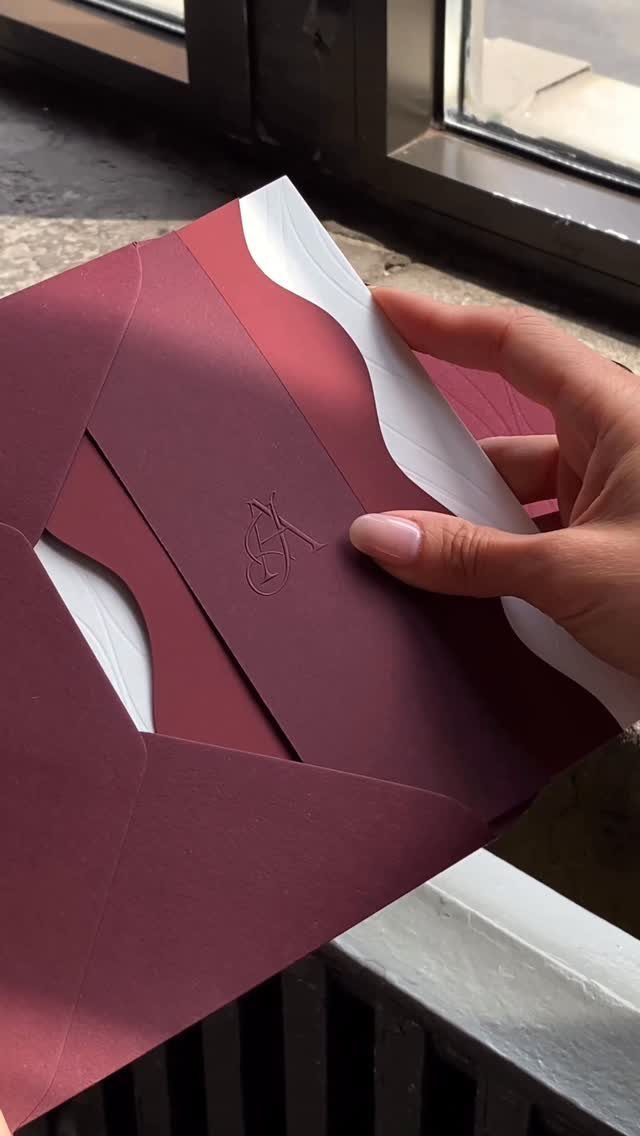 Burgundy wedding invitation suite with a white wavy insert held by a hand.