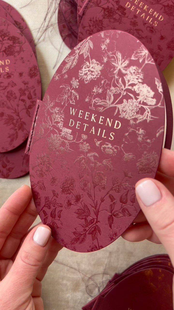 Burgundy oval wedding detail cards with floral patterns held by two hands.