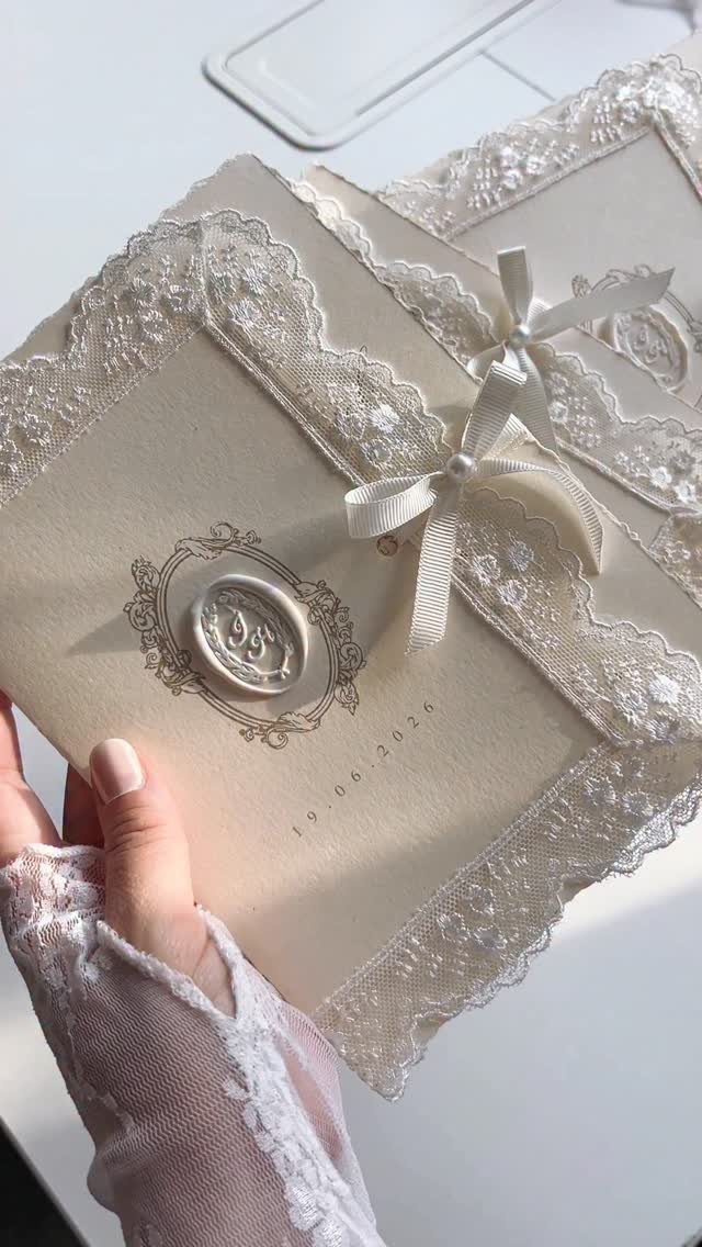 Cream wedding invitation with white lace, a satin bow, and a wax seal.