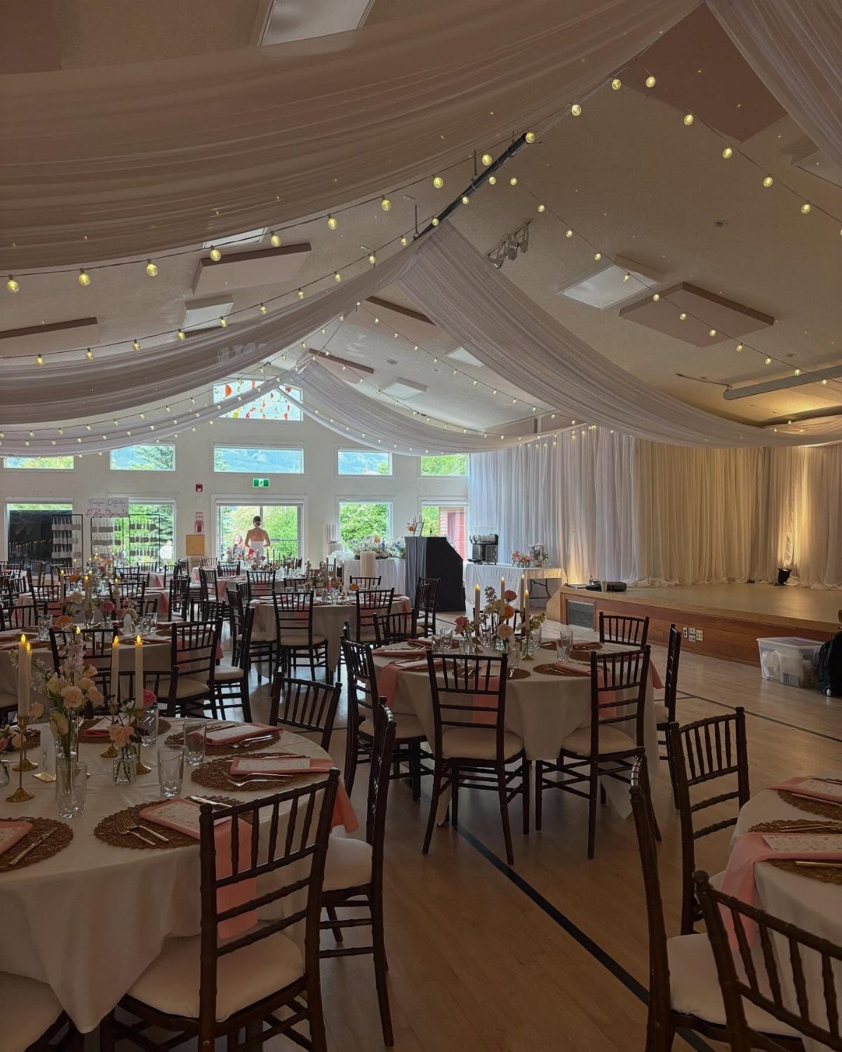Wedding reception in a hall with white ceiling draping, string lights, and set tables.