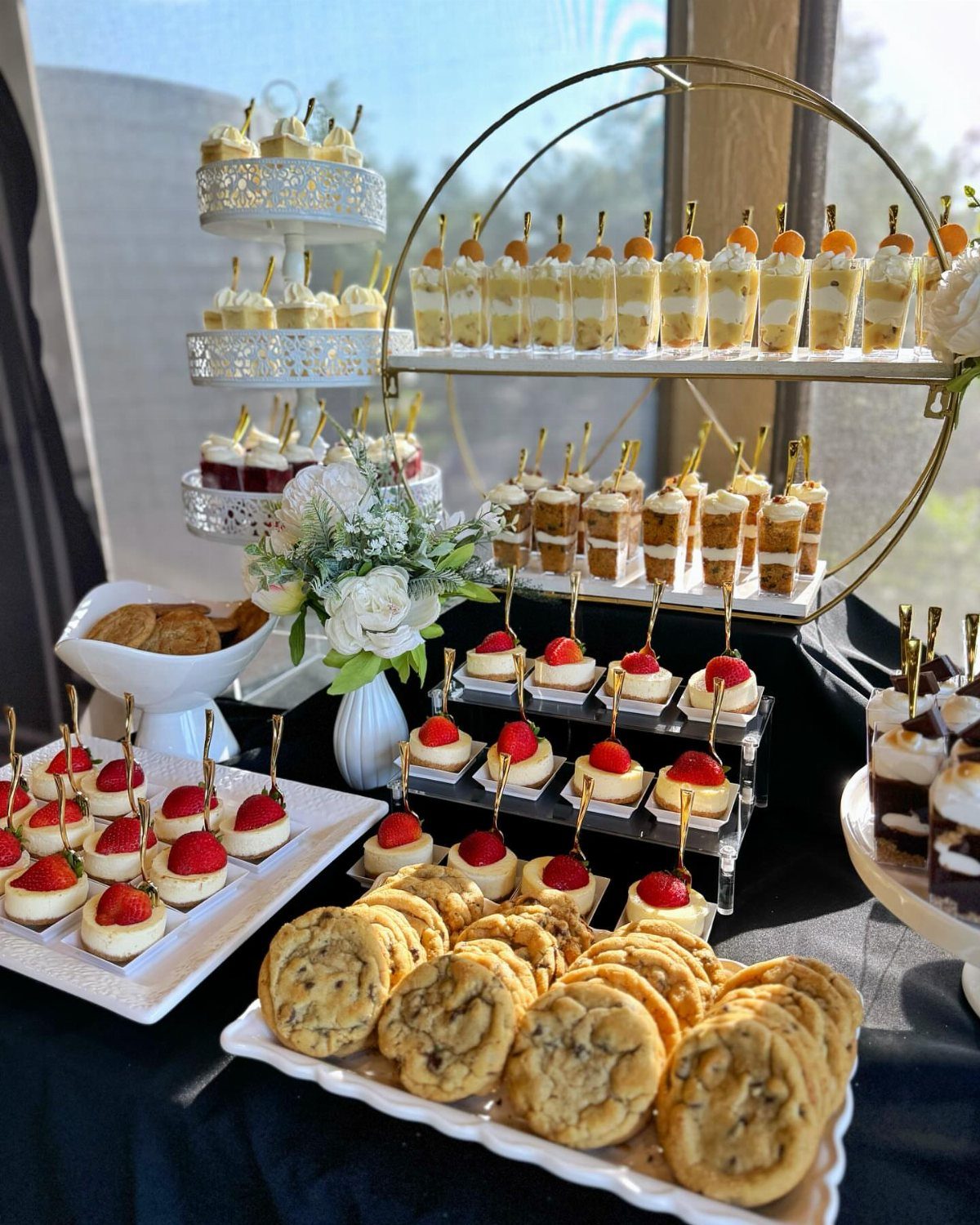 Dessert bar with cookies, mini cheesecakes, and parfaits on a black table.