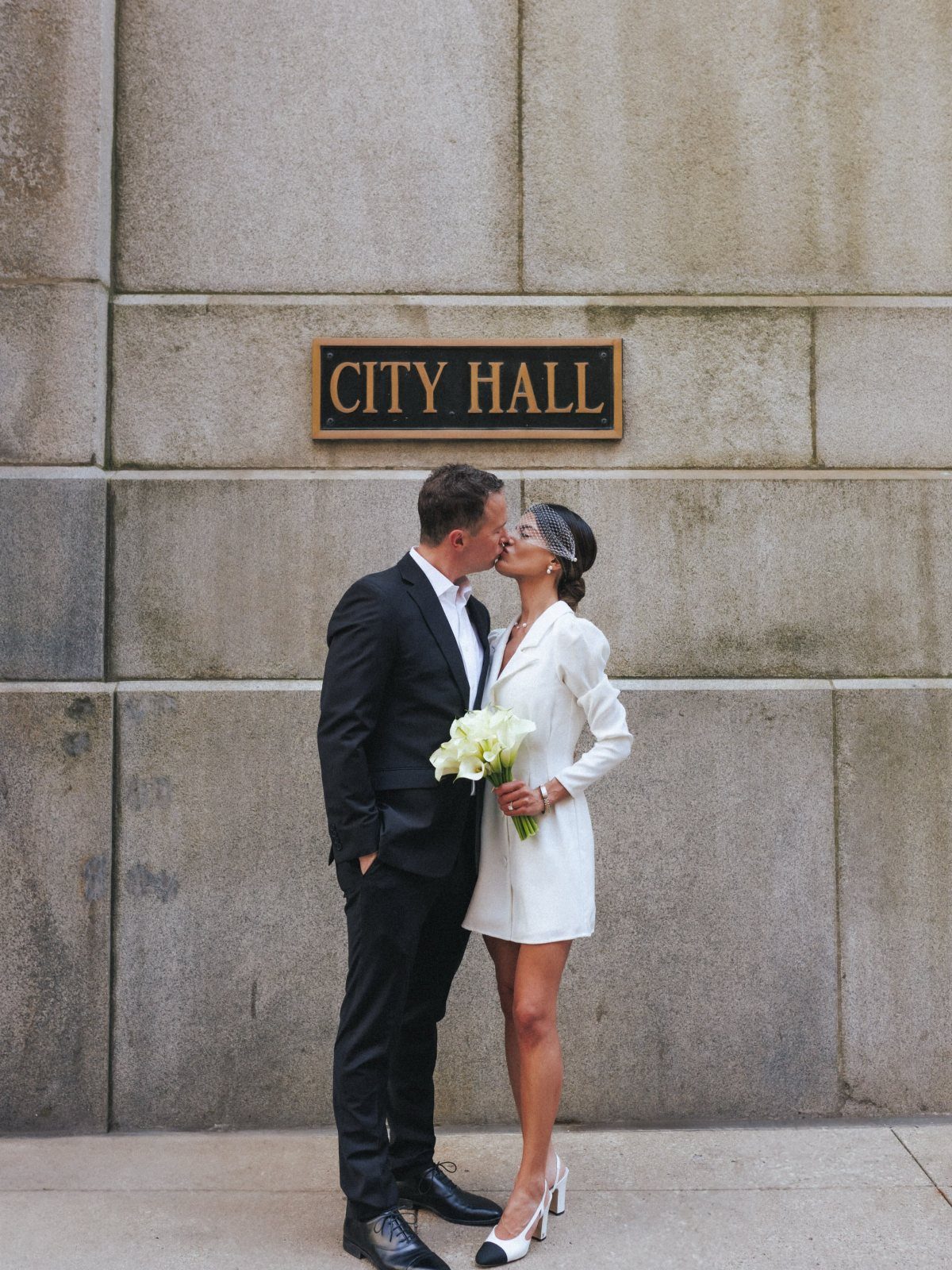Couple kissing in front of City Hall, woman in short white dress holding bouquet.