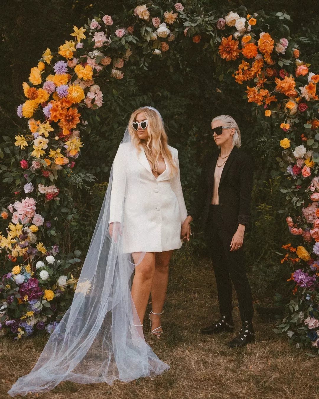 Two brides standing under a vibrant floral arch in an outdoor park setting.