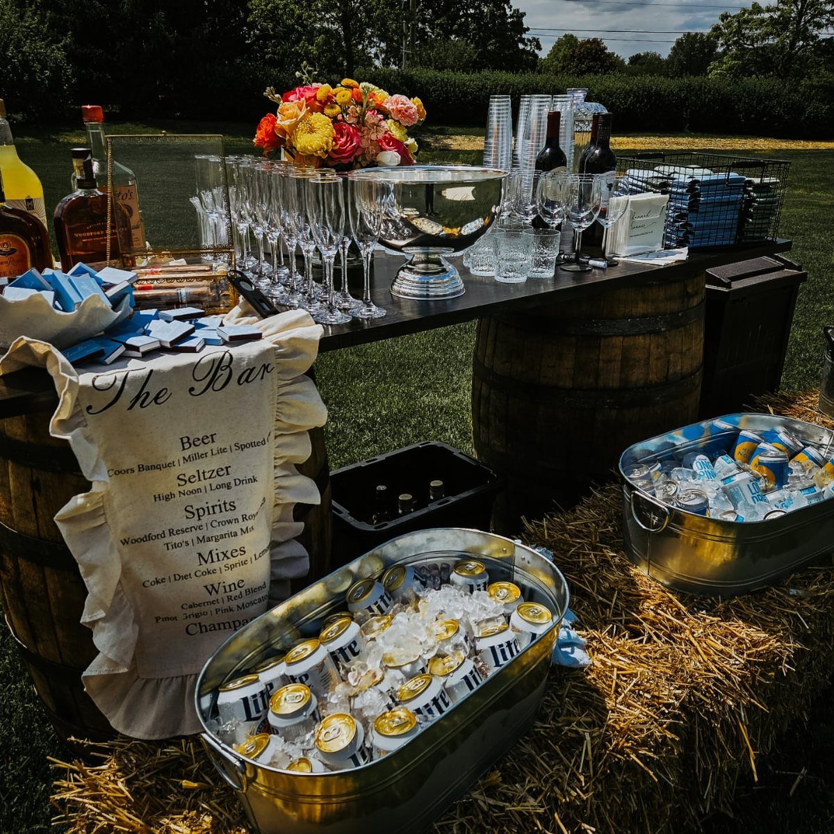 Outdoor self-serve wedding bar with drinks in ice tubs on wooden barrels and hay.