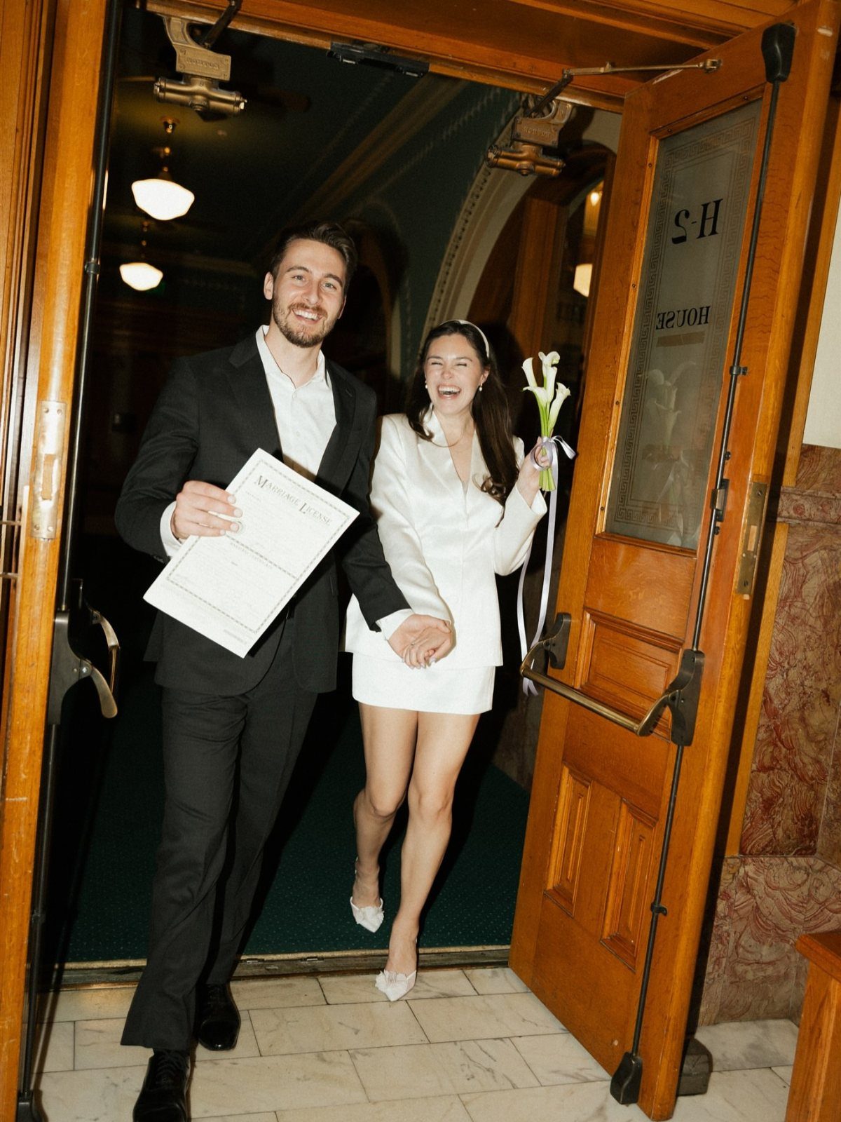 Newlywed couple walking through wooden doors, man in suit and woman in short dress.