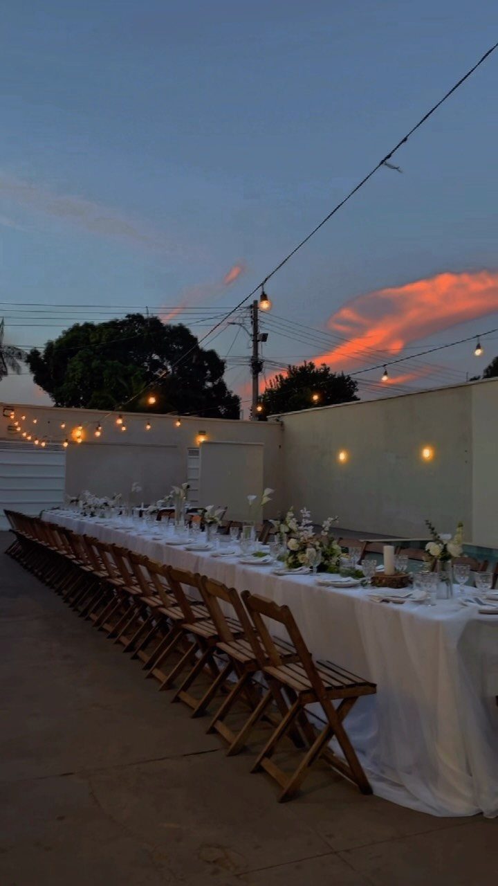 Long wedding table with white linens and wooden chairs under glowing string lights.