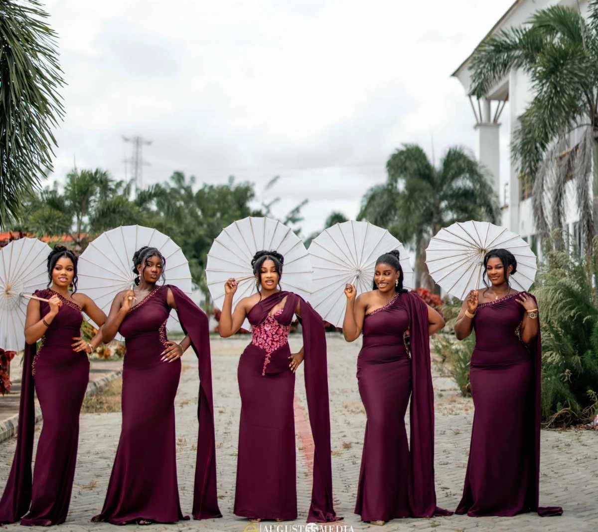 Five bridesmaids in burgundy one-shoulder gowns holding white parasols on a garden path.