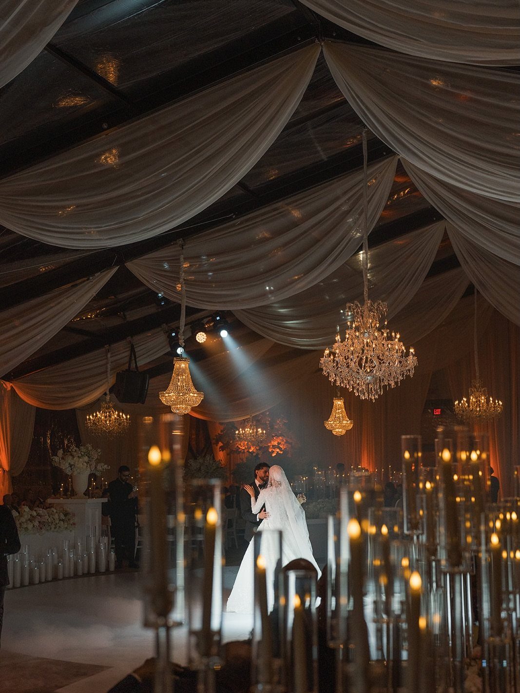 Bride and groom kissing under crystal chandeliers and white fabric ceiling with candles.