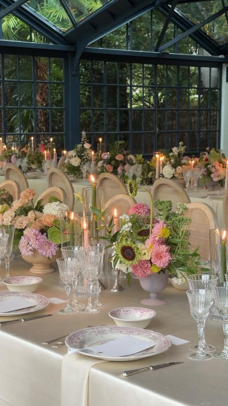 Elegant dining table with floral centerpieces and candles in a glass conservatory.