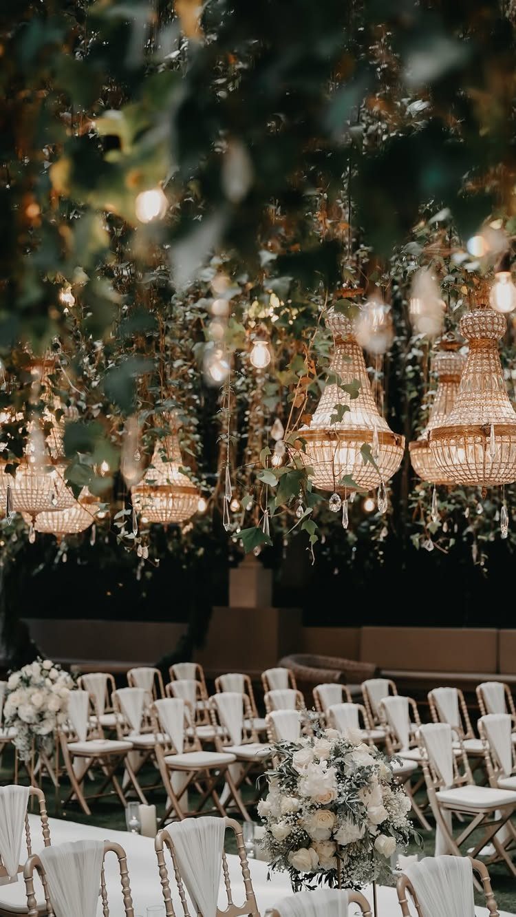 Wedding ceremony setup with white chairs under a canopy of greenery and chandeliers.