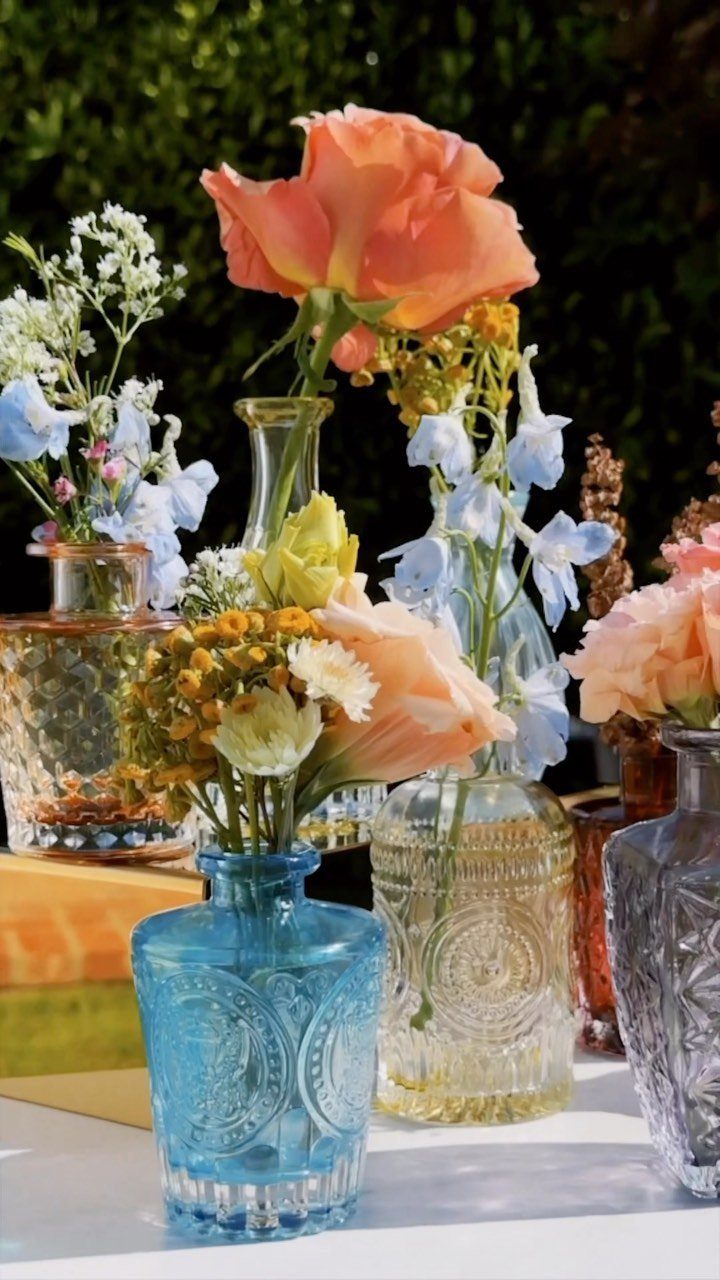 Colorful glass vases with fresh orange and blue flowers arranged outdoors on white.