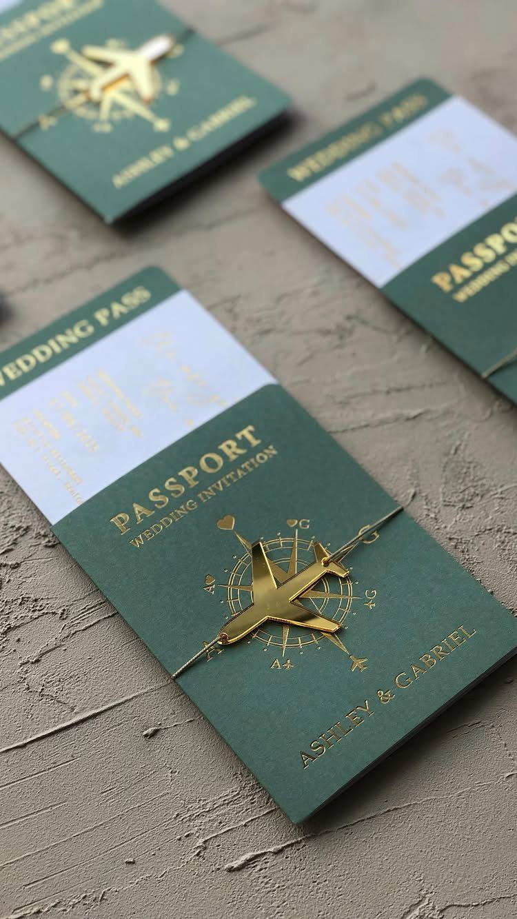 Green passport-style wedding invitations with gold airplane charms on a beige surface.