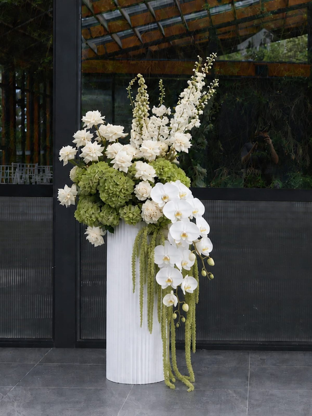 Large white and green floral arrangement on a white fluted pedestal indoors.