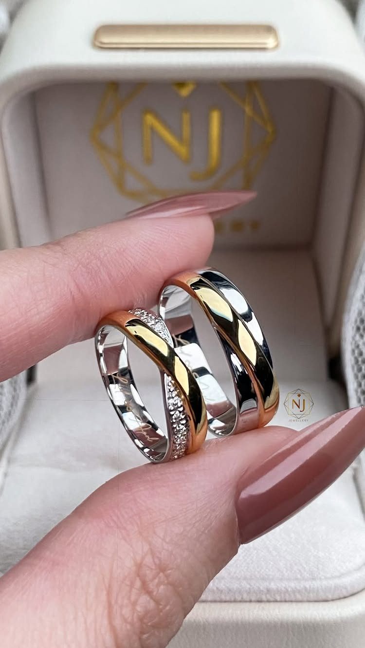 Two gold and silver wedding bands held by fingers over a white jewelry box.