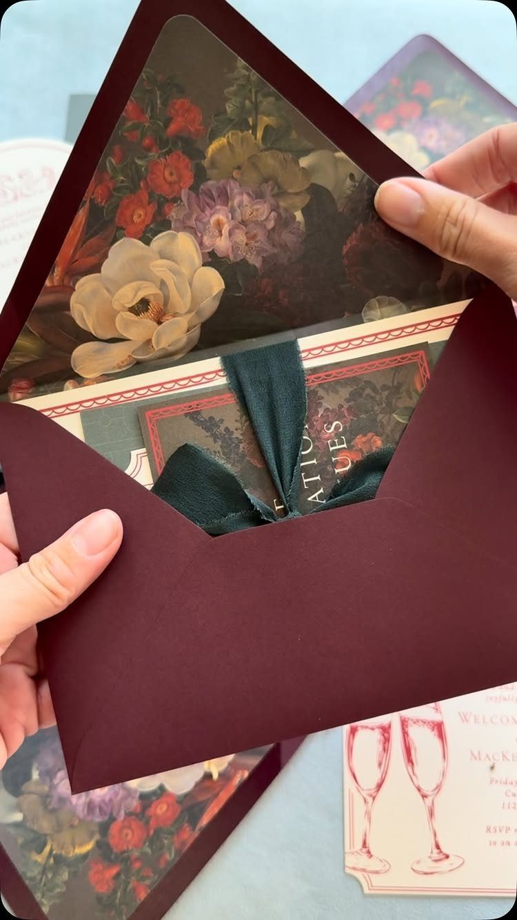 Hands opening a burgundy envelope containing floral wedding invitations tied with green ribbon.