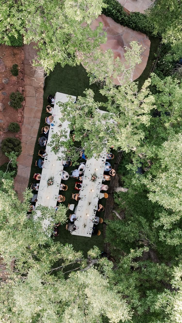 Long white dining tables set outdoors with guests seated under green trees.