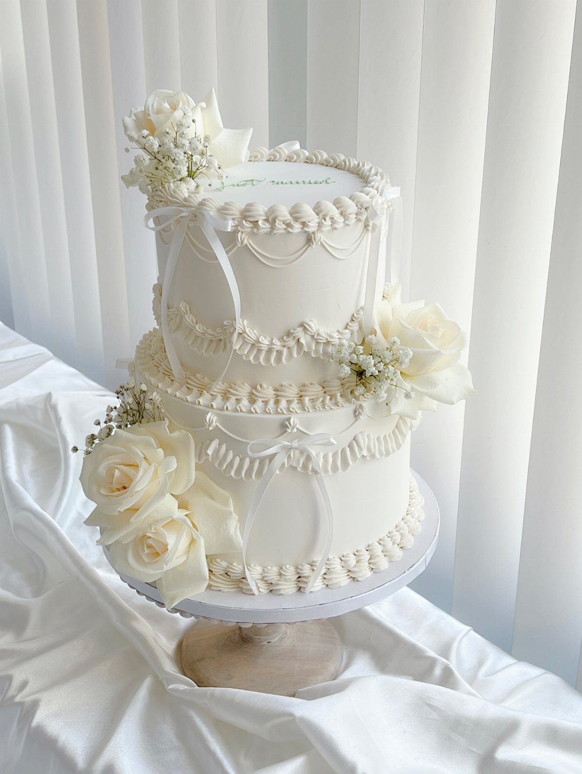 Two-tier white wedding cake decorated with white roses and ribbons on a stand.
