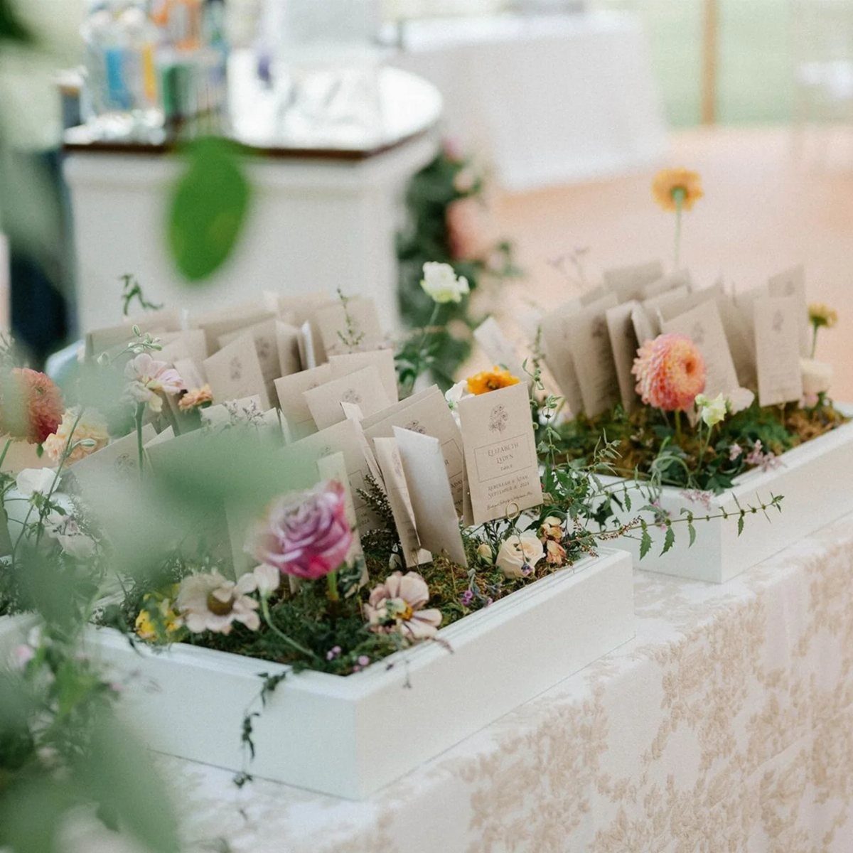 Brown paper favor envelopes nestled in white planters with colorful spring flowers.
