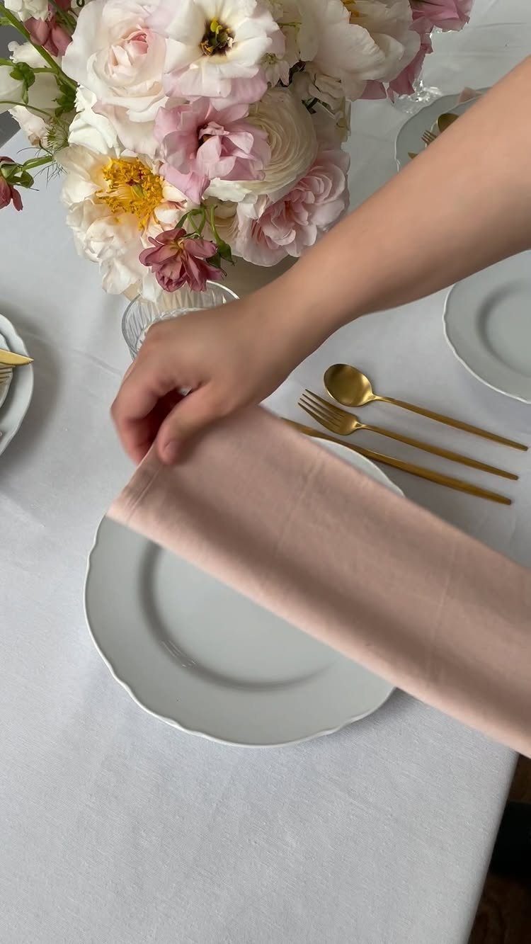 Hand placing a pink napkin on a white plate with gold cutlery.