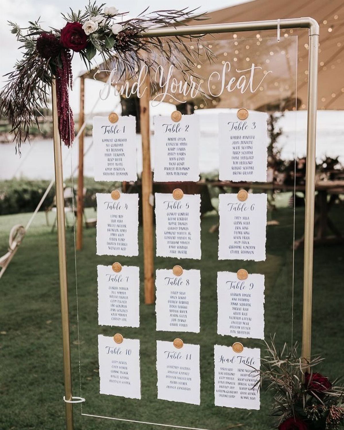 Acrylic wedding seating chart with white guest lists on a gold stand outdoors.