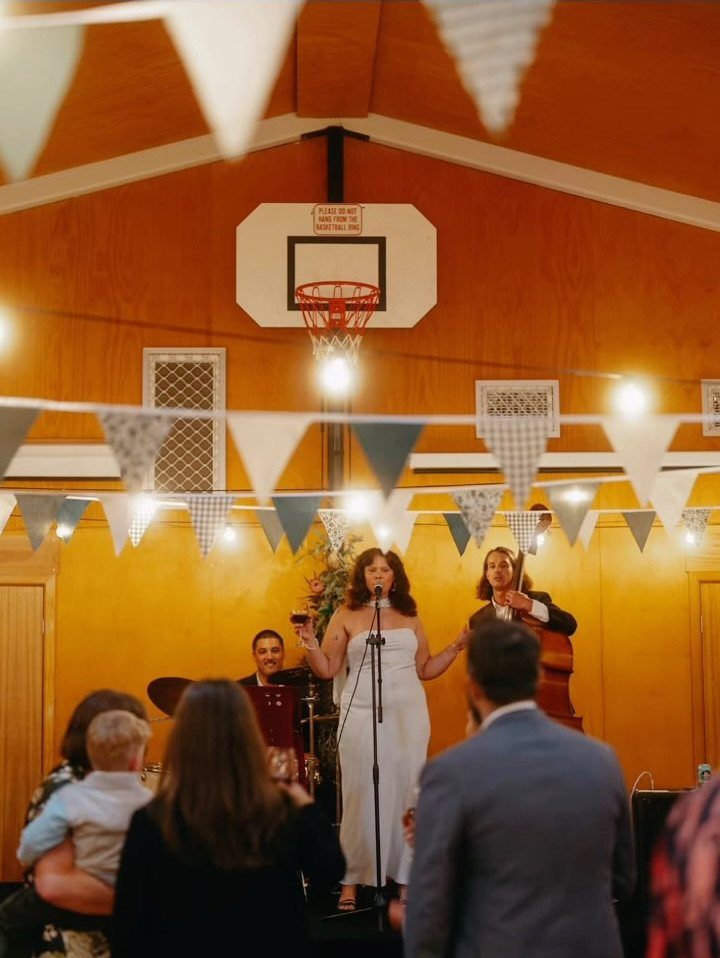 Bride speaking at a wedding reception in a community hall with a basketball hoop.
