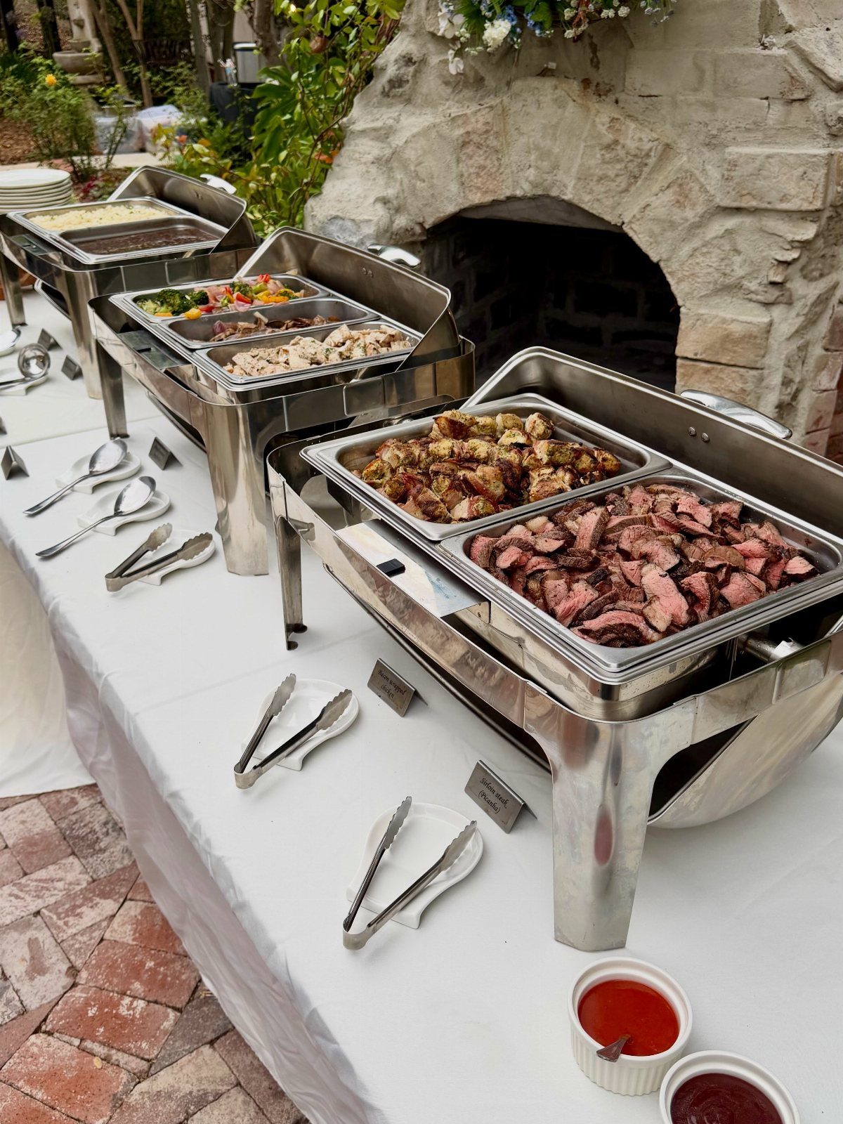 Buffet style food in stainless steel chafing dishes on a white tablecloth outdoors.