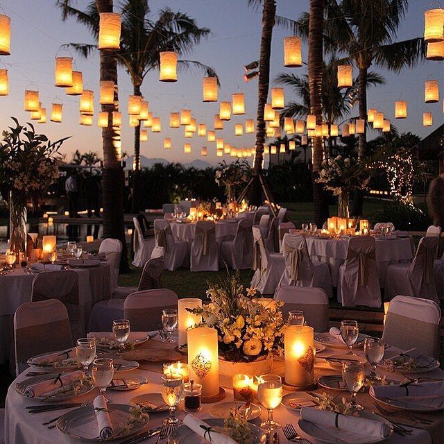 Outdoor wedding reception tables with white linens, candles, and hanging paper lanterns at dusk.