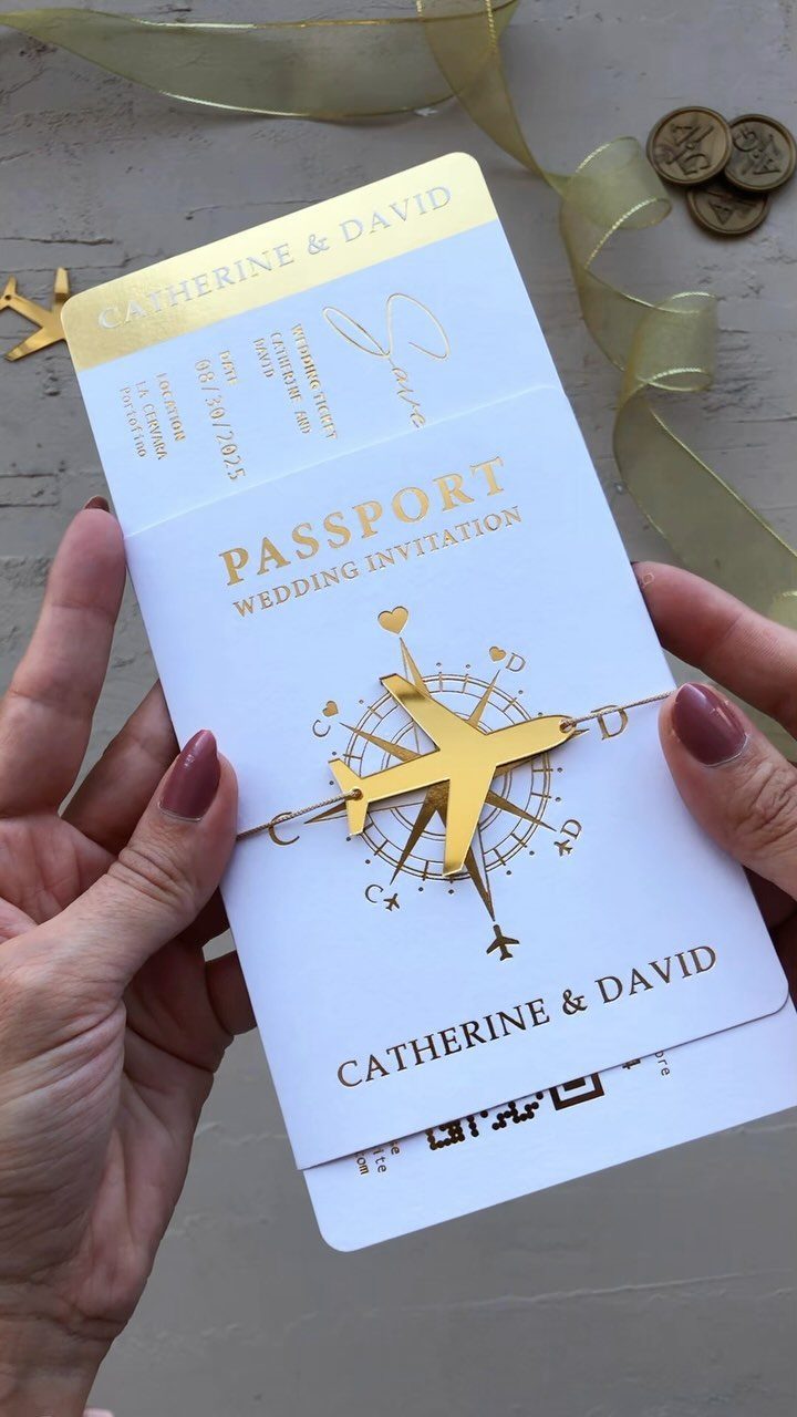 Passport-style wedding invitation with gold foil airplane and compass held by two hands.