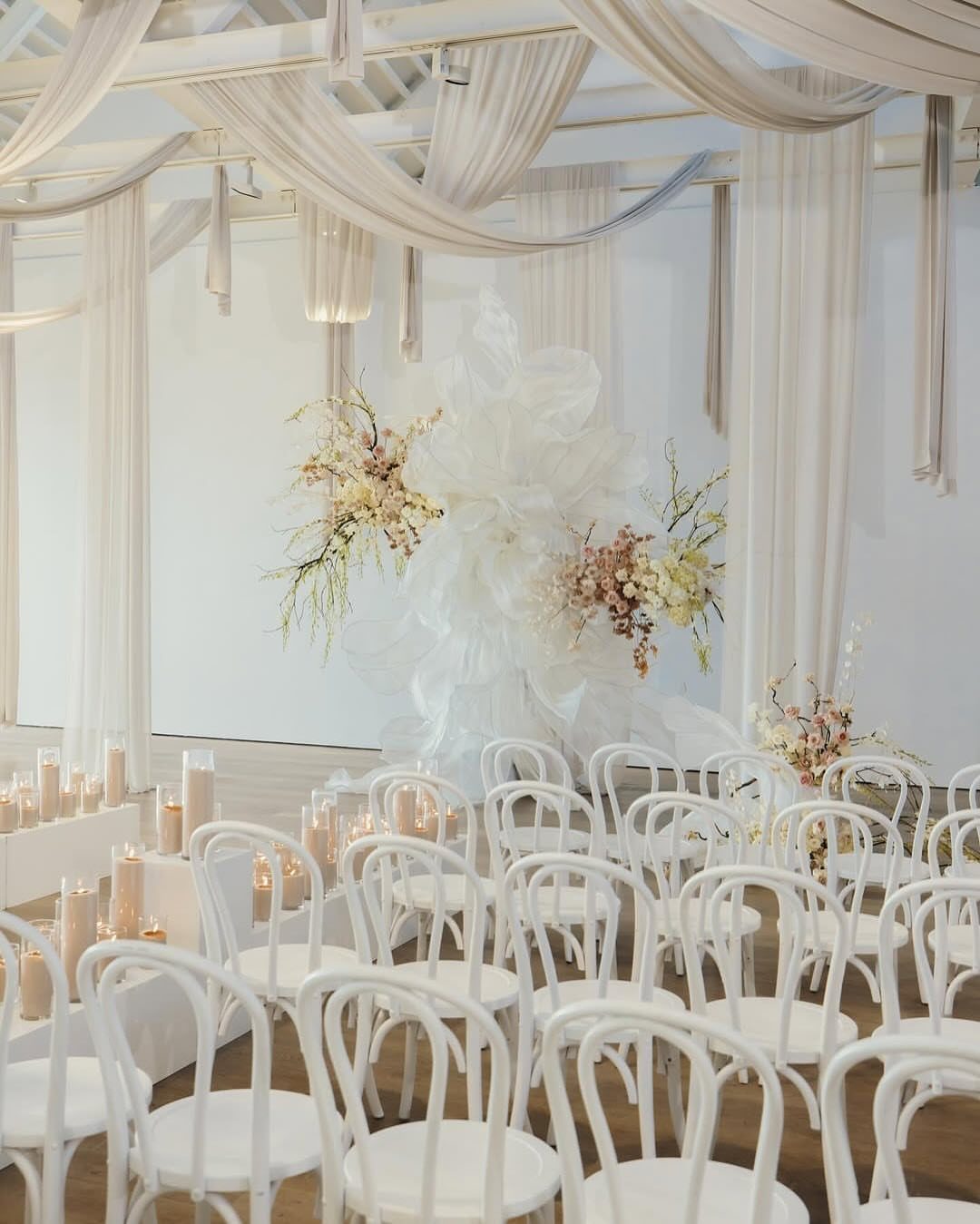 White wedding ceremony setup with bentwood chairs, fabric draping, and white floral arrangements.