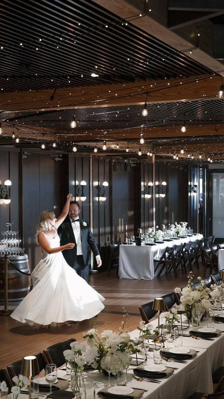 Bride and groom dancing in a modern wedding venue with string lights and tables.