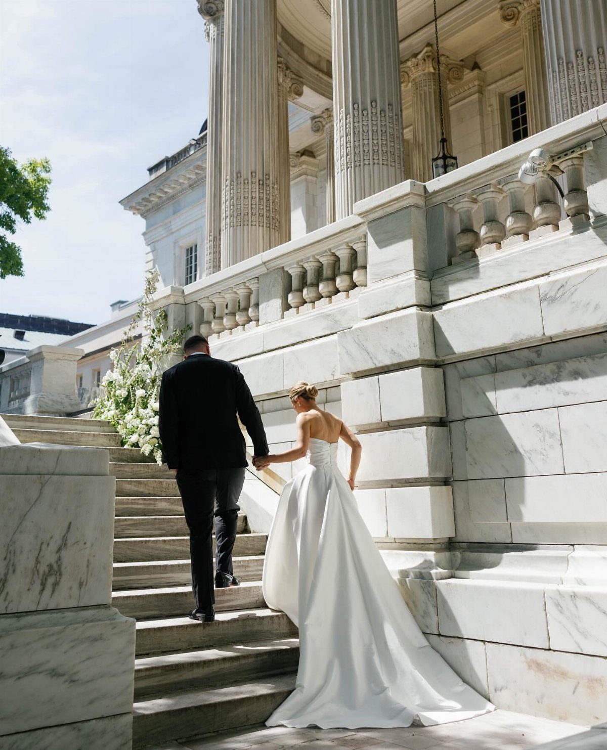 Bride and groom walking up white marble stairs toward a classical columned portico.