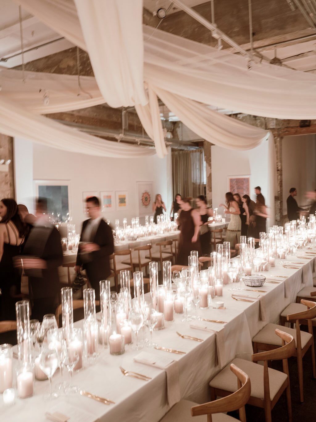 Long banquet table with white linens and many lit candles in an industrial venue.