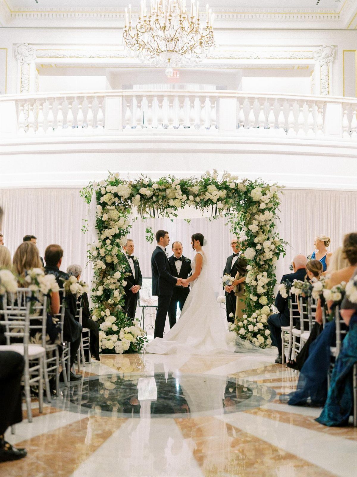 Wedding couple under a floral arch in a grand hall with marble floors.