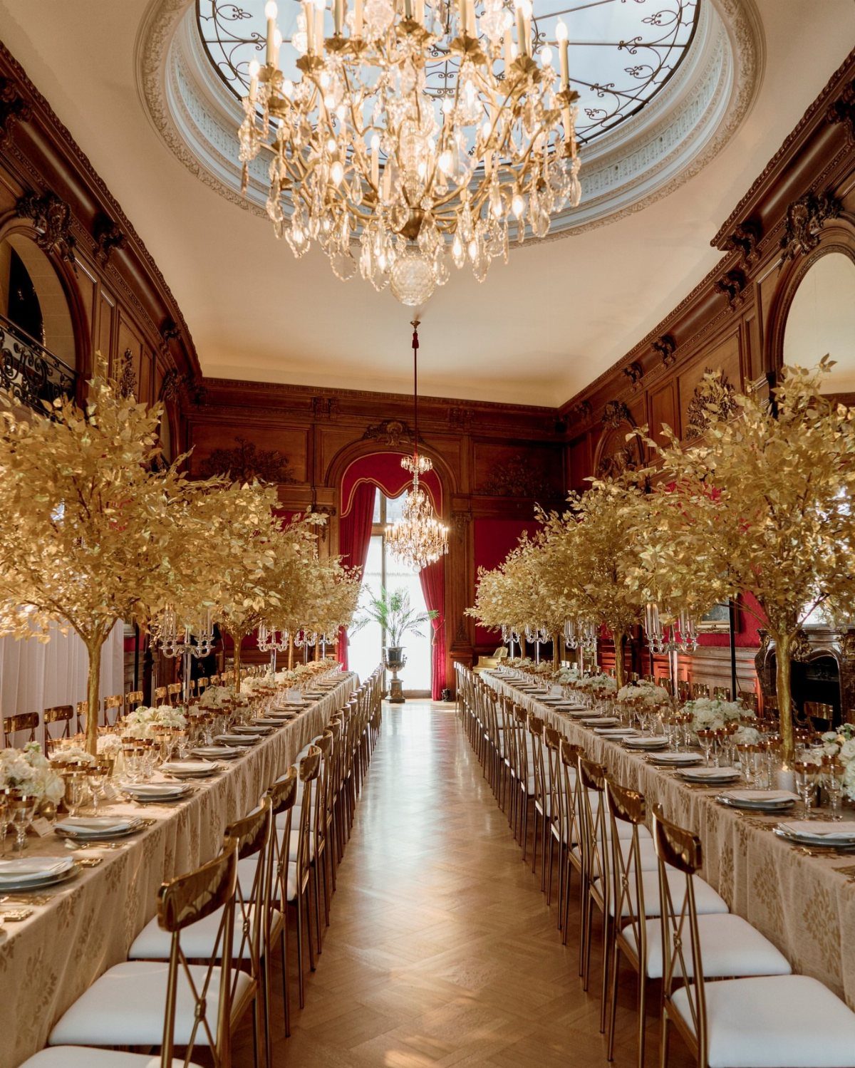 Opulent banquet room with long tables, gold chairs, and crystal chandeliers under a dome.