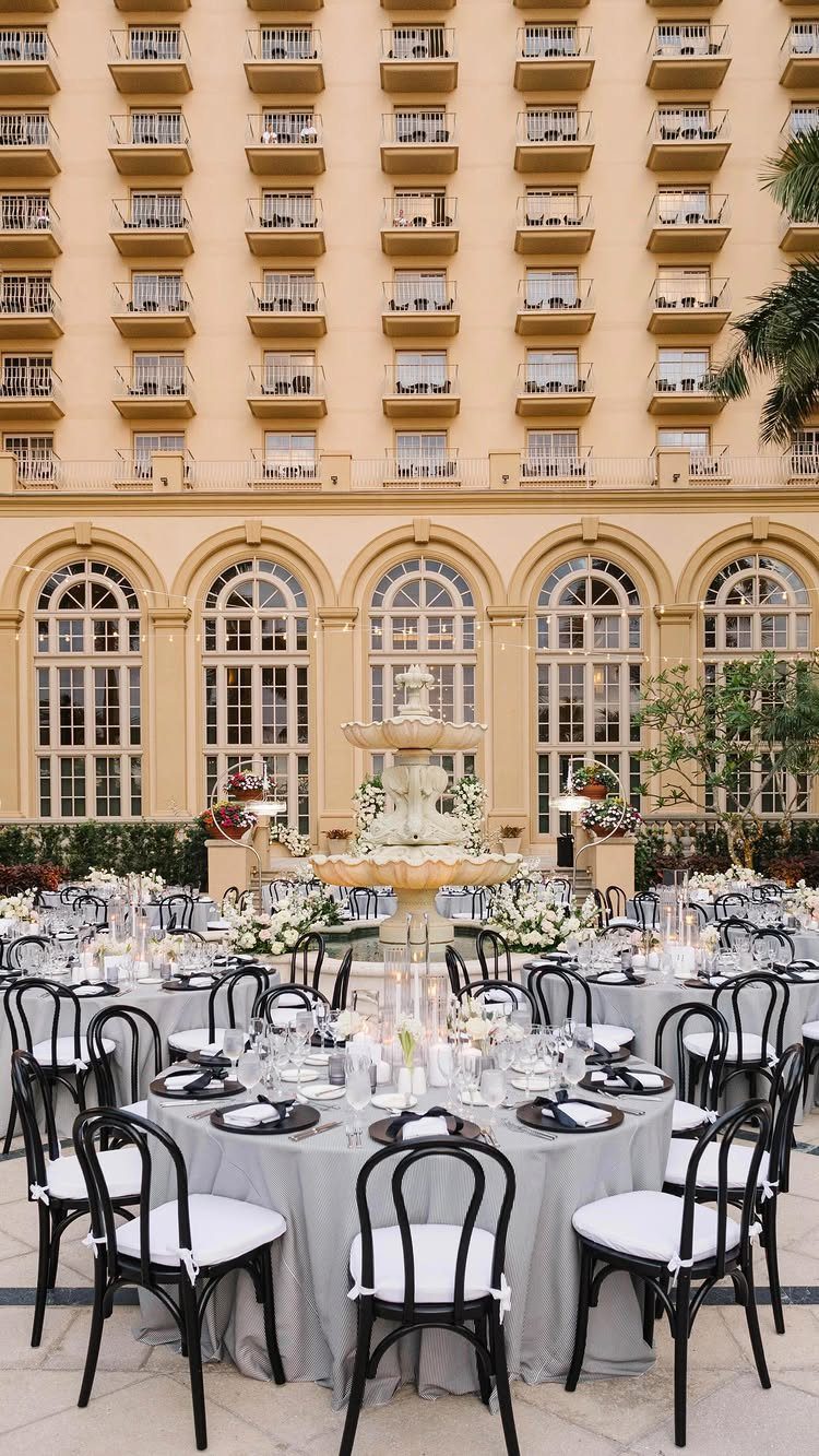 Formal outdoor courtyard event setup with grey tables, black chairs, and a stone fountain.