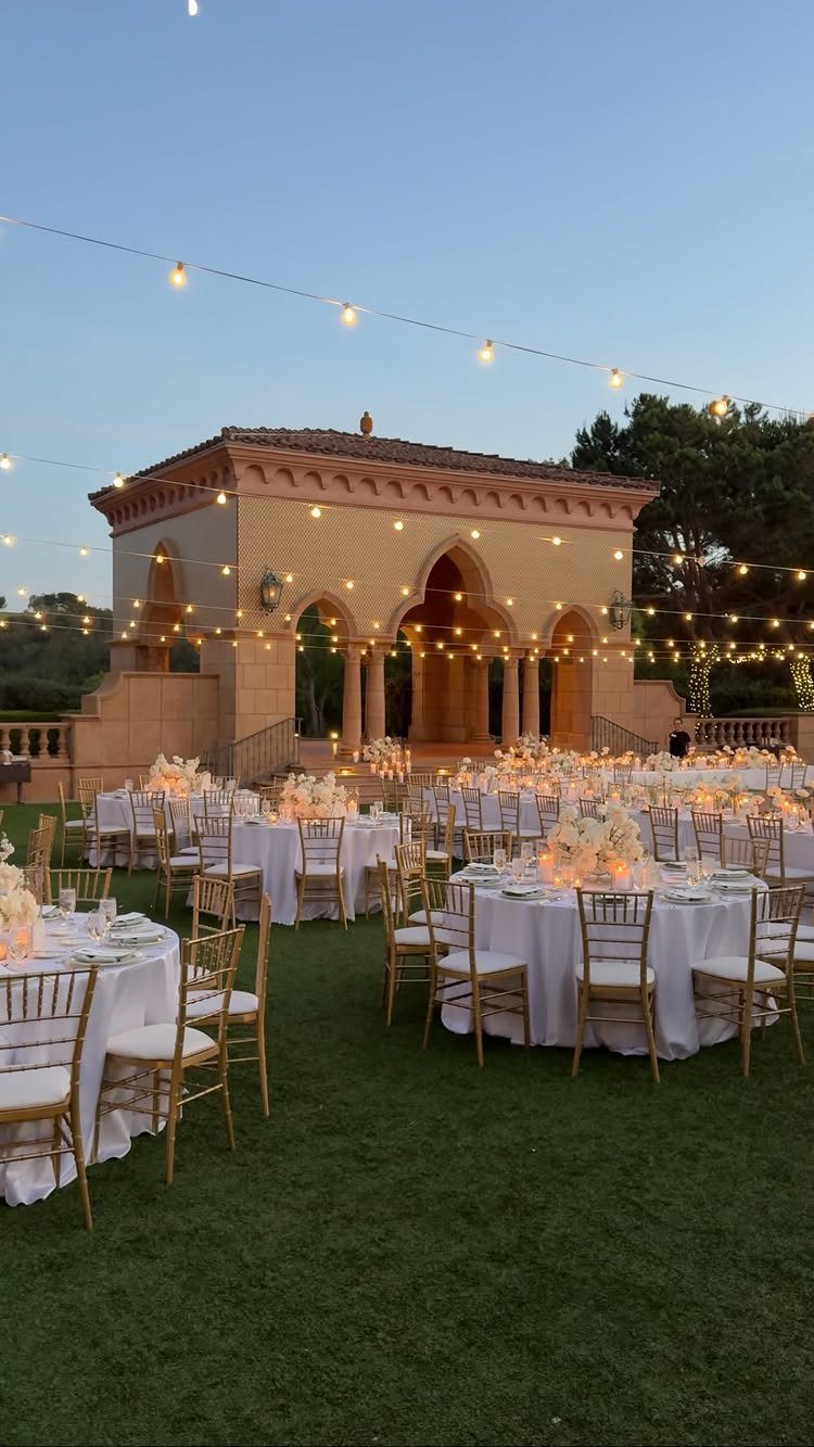 Outdoor wedding reception with white tables and gold chairs before a stone pavilion.