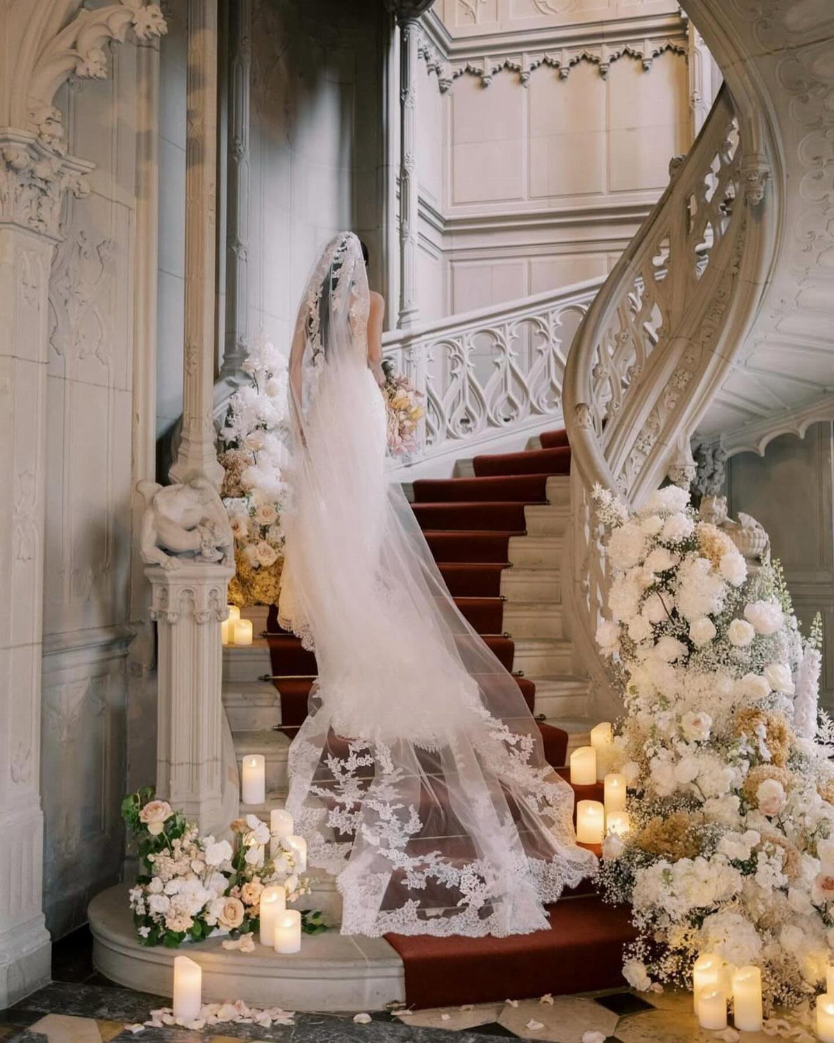 Bride in white gown standing on a grand staircase with red carpet and flowers