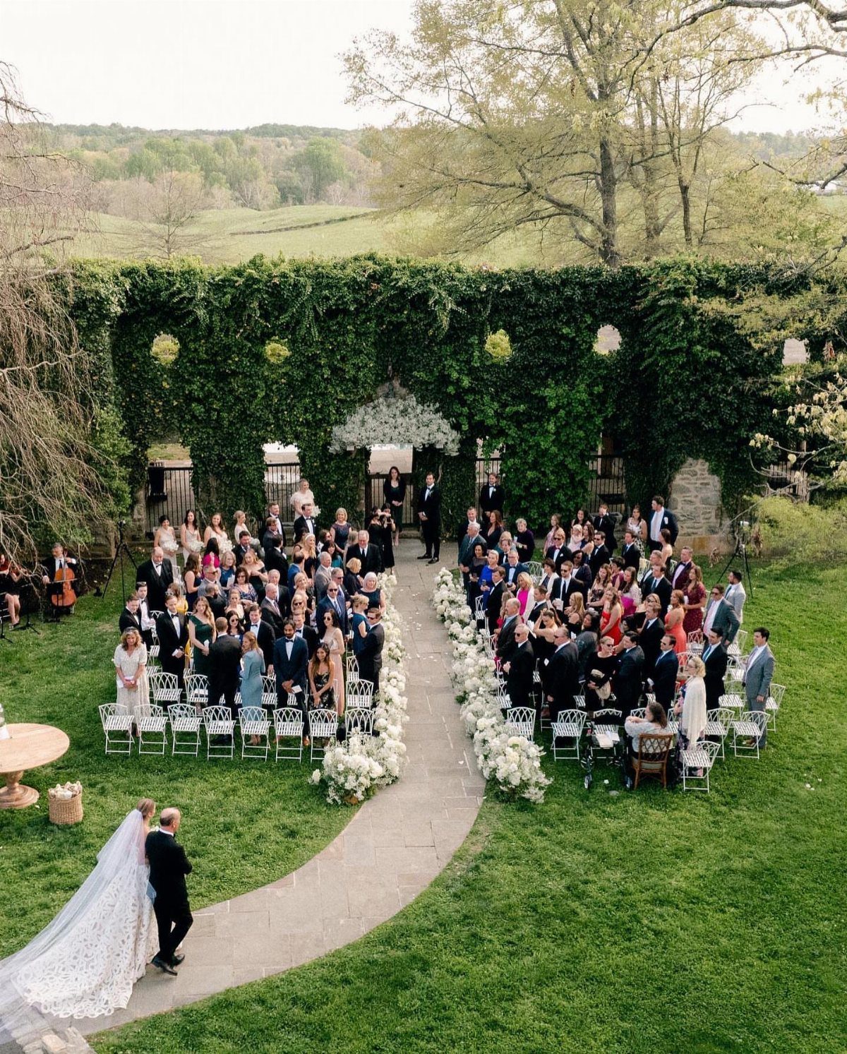 Outdoor wedding ceremony with guests seated along a flower-lined path in a lush garden.