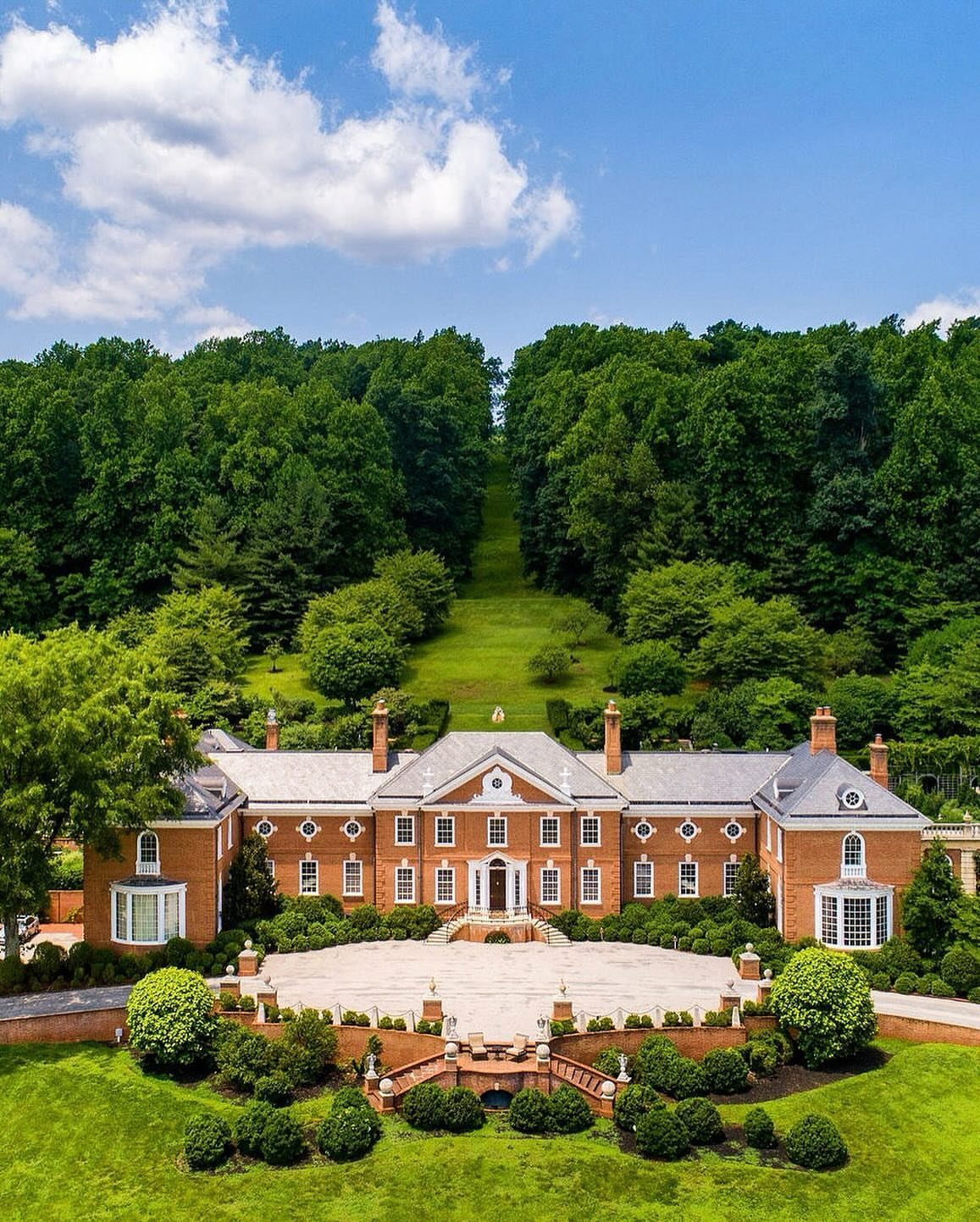 Large red brick mansion with a stone patio surrounded by lush green forests.