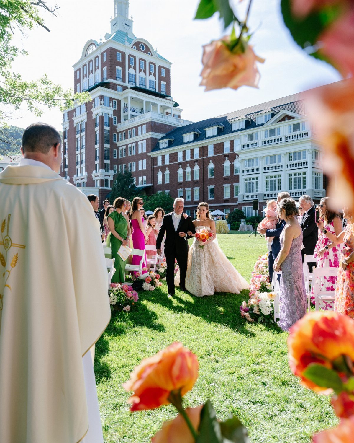 Bride and groom walking down a grassy aisle toward a priest at a resort.
