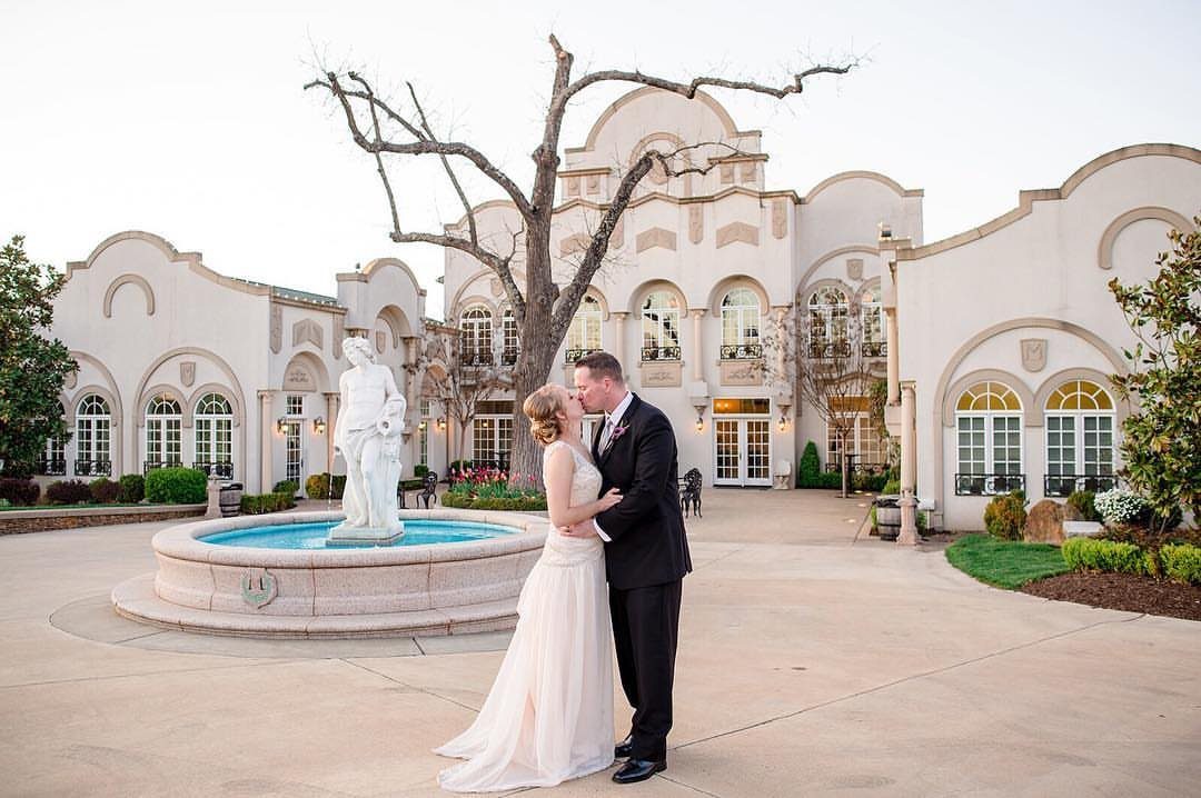 Bride and groom kissing before a grand white estate with a fountain statue.