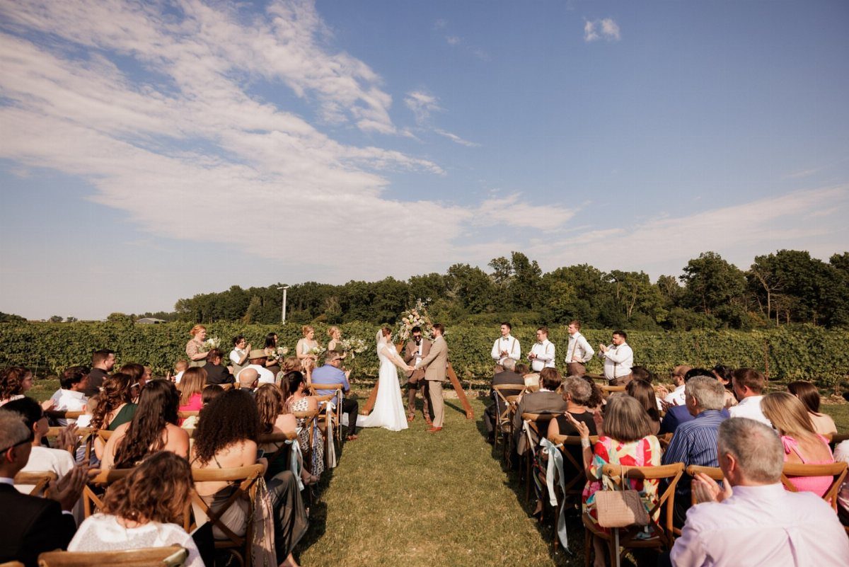 Wedding ceremony taking place in a vineyard with guests seated on a lawn.