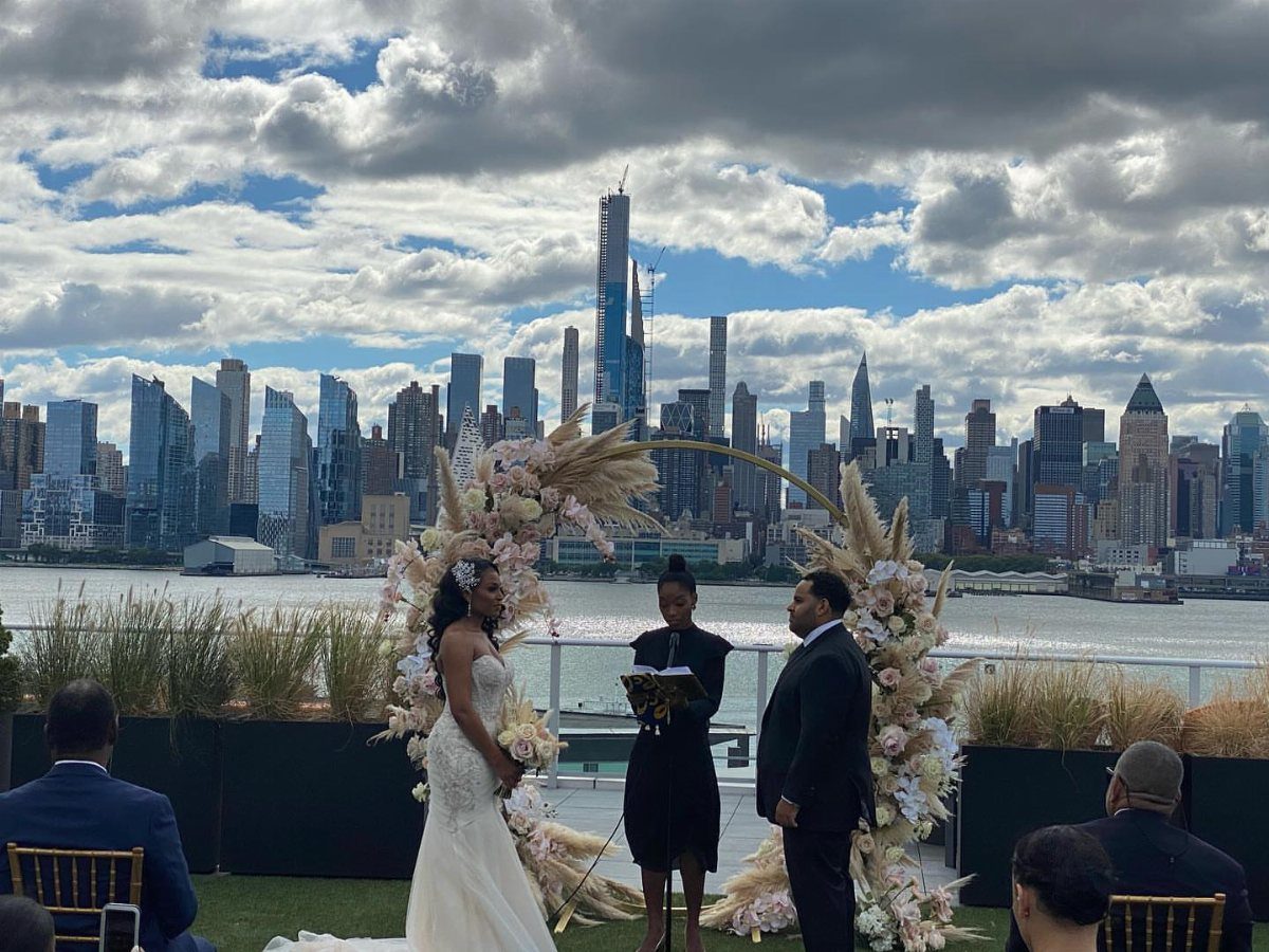 Wedding couple and officiant under a floral arch with Manhattan skyline background.