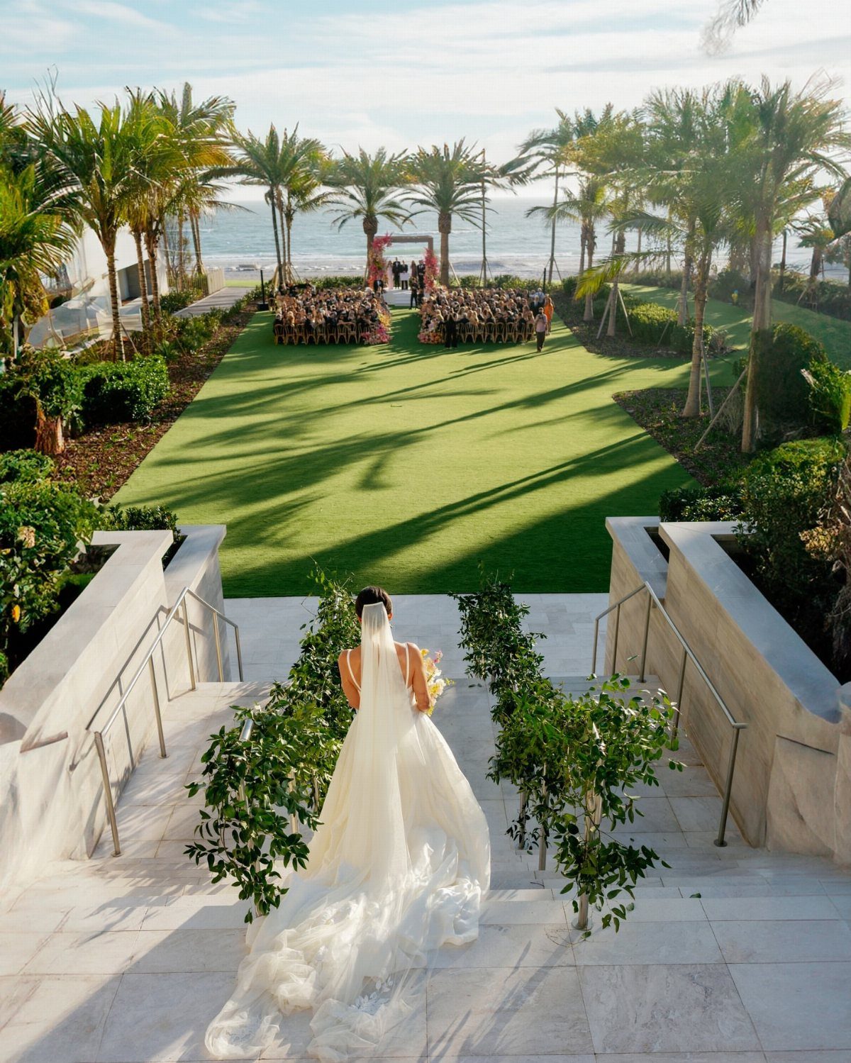Bride walking down white stairs toward a beach wedding ceremony with palm trees.