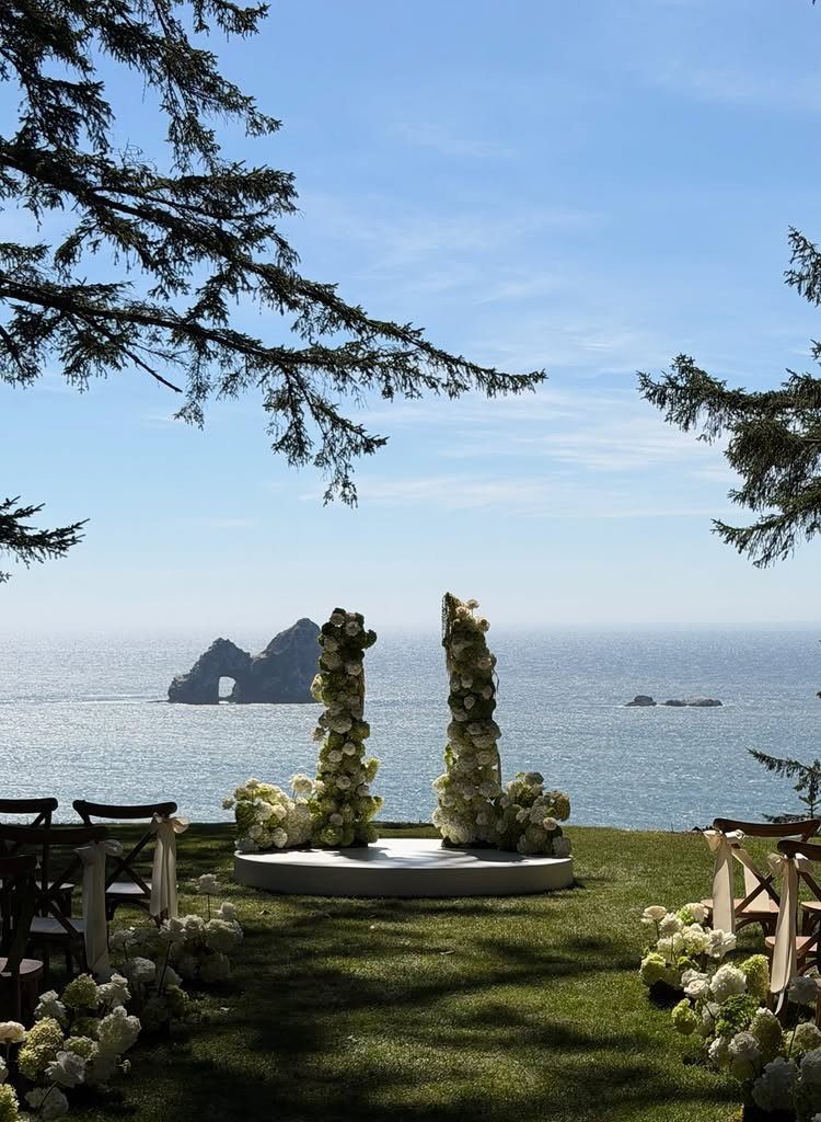 Wedding ceremony setup with white floral pillars on a cliff overlooking the ocean.